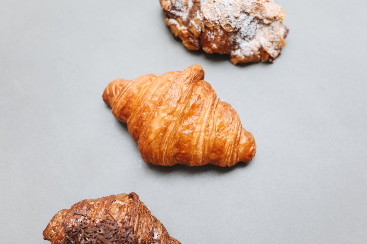 A variety of croissants including plain, chocolate, and powdered sugar on a gray surface.